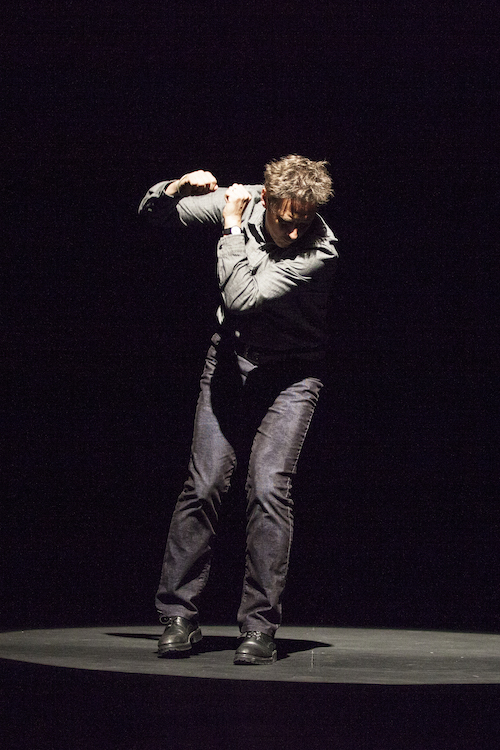 A man in jeans and button up holds his fists up by this shoulders under a spotlight.